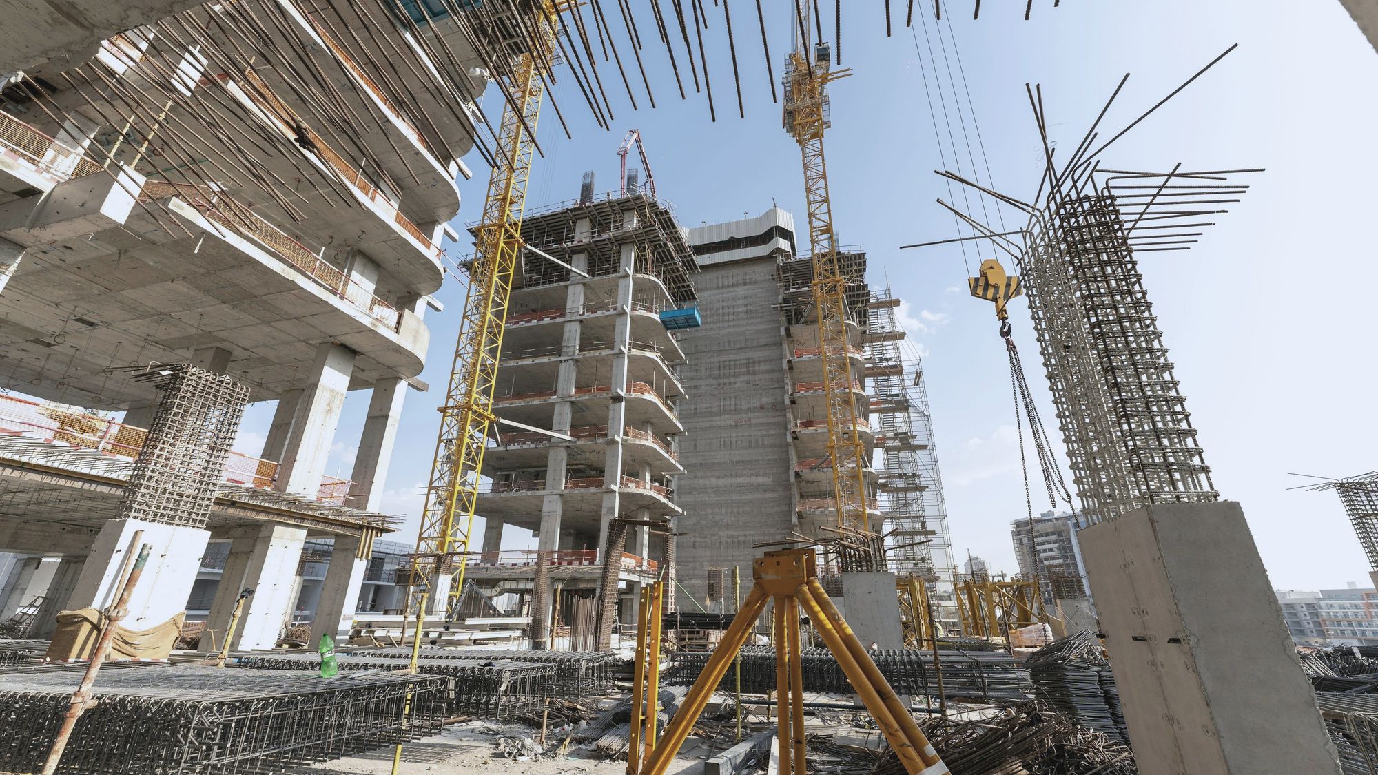 A concrete building rising among tower cranes, seen from below beside a survey tripod