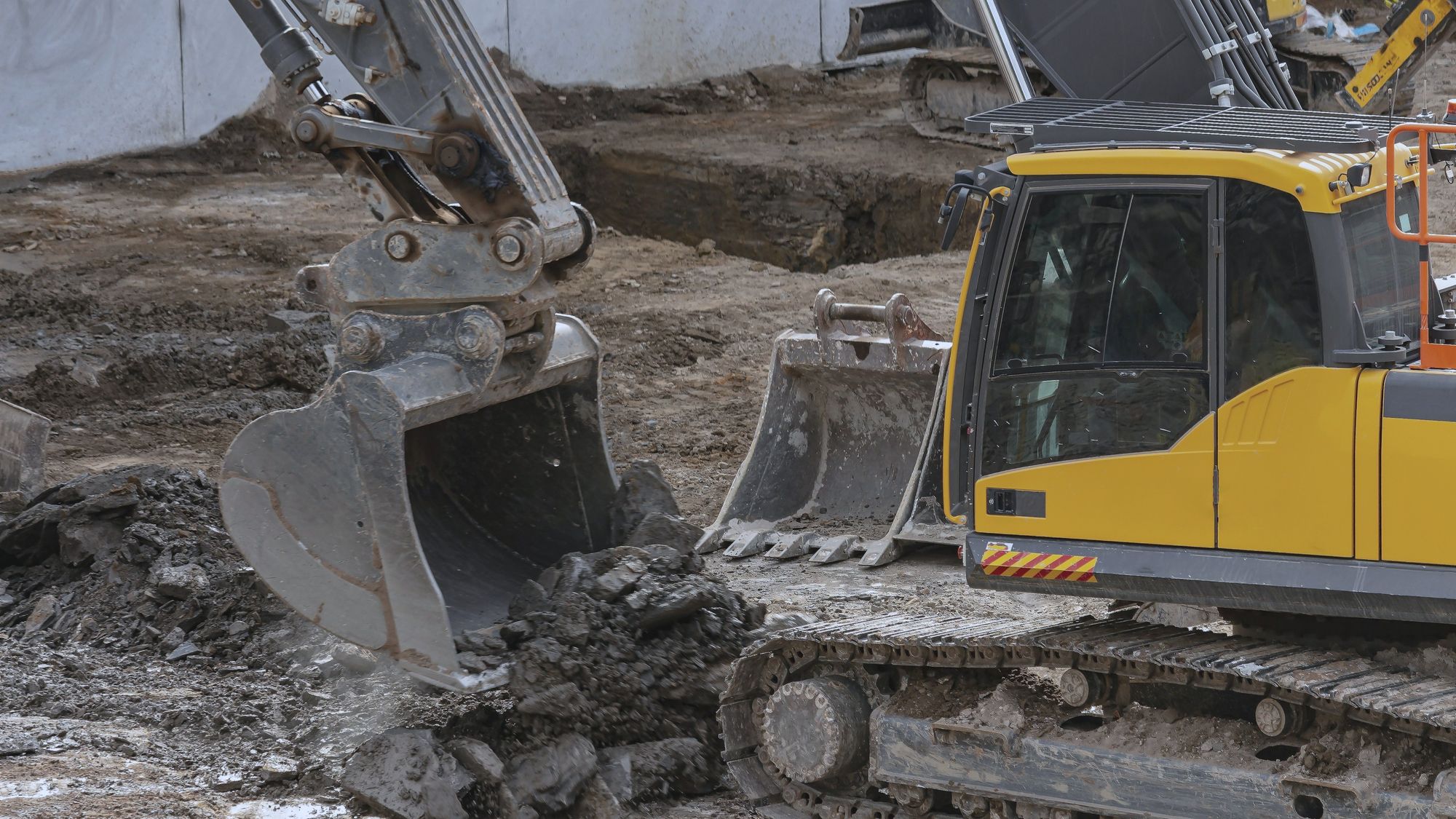 Two excavators digging into a muddy excavation, buckets full of soil